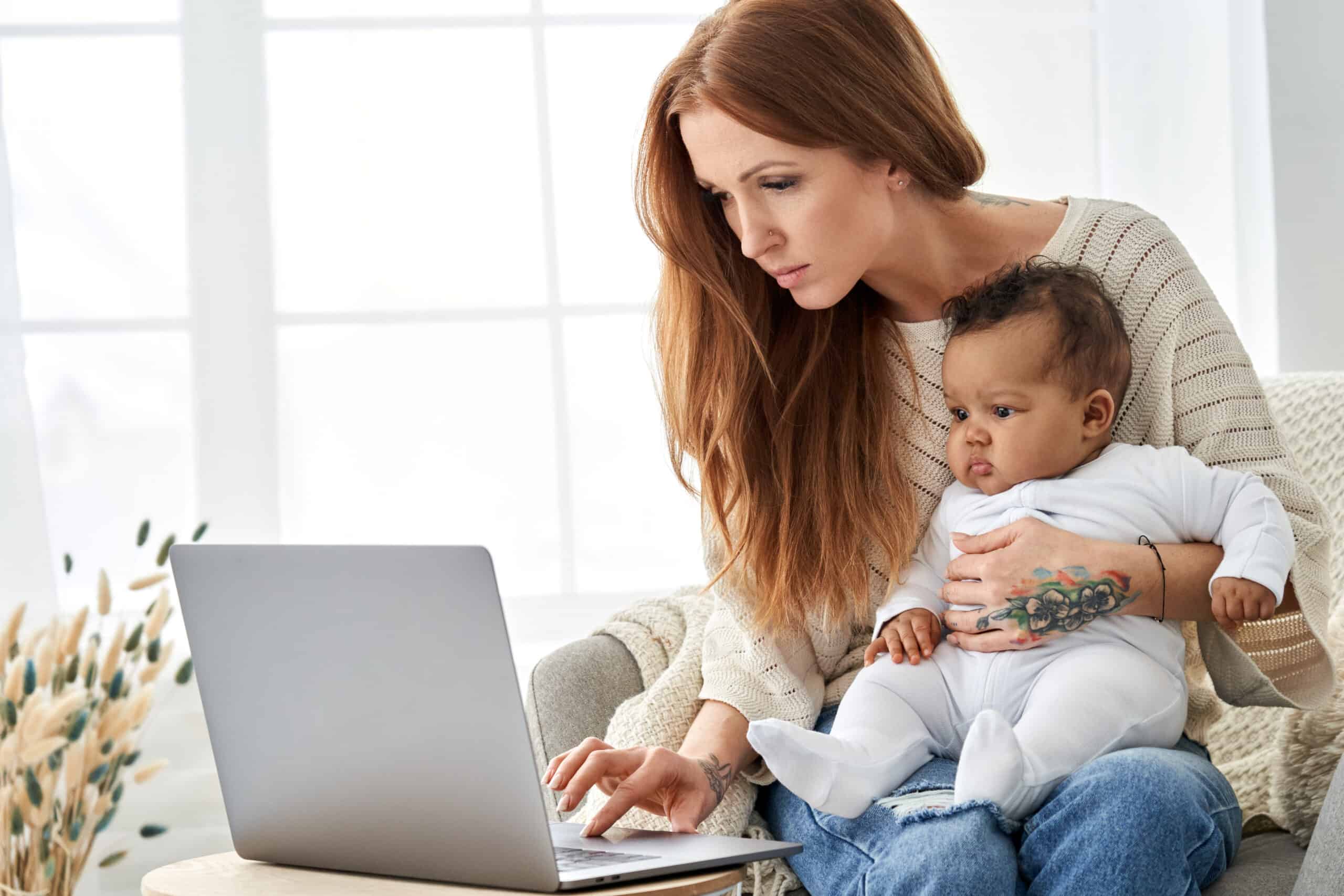  Busy mother using laptop working from home with little baby.